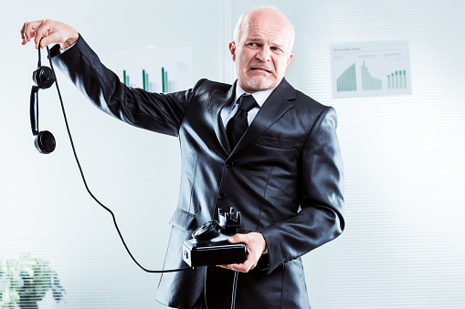 Upset businessman holding a telephone at arms length dangling the receiver from his fingertips with a grimace