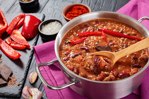 hot delicious chili con carne with whole red hot chilis, kidney beans, tomatoes and piece of chocolate in a pot with ingredients at background, authentic recipe, view from above, close-up