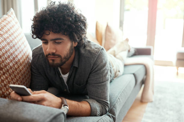 Chilling on my free time Shot of a handsome young man using his cellphone while relaxing on the couch at home scrolling photos stock pictures, royalty-free photos & images