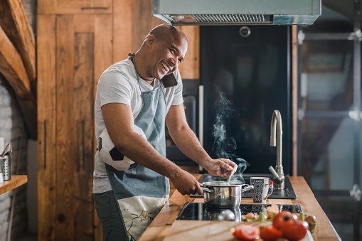 Happy black man holding soccer ball while communicating on cell phone and preparing food in the kitchen.