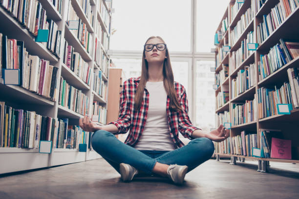 refrigeração, bem-estar, vitalidade, paz, sabedoria, educação, estilo de vida do campus. tiro de ângulo baixo de jovem garota nerd calma, praticando yoga em posição de lótus no chão na sala de arquivo de biblioteca - balance book university glasses - fotografias e filmes do acervo