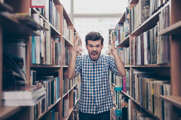 istock Going crazy and insane, psycho and agression. Shouting stressed young brunet bearded student is blasting standing between the book shelves in the library 935978580
