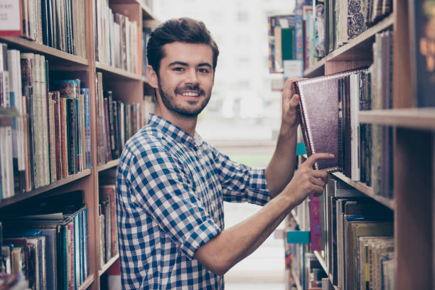istock Cheerful clever young attractive brunet bearded student bookworm is studying in the ancient university library, putting vocabulary on the book shelf, smiling, wearing casual checkered shirt 935982662