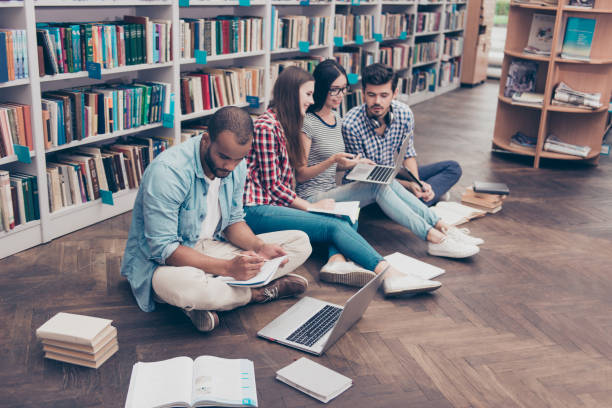 vier fröhliche konzentriertere internationalen klassenkameraden sitzt auf dem holzboden in der alten bibliothek auf dem campus und studie, prüfungsvorbereitung, tests, mit büchern, notizblöcke, laptops - nerd computer learning fun stock-fotos und bilder