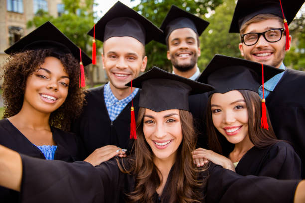 selfie for memories. six with cheerful graduates are posing for selfie shot, attractive brunette lady is taking, wearing gowns and mortar boards, outside on a summer day - graduation student women beauty imagens e fotografias de stock