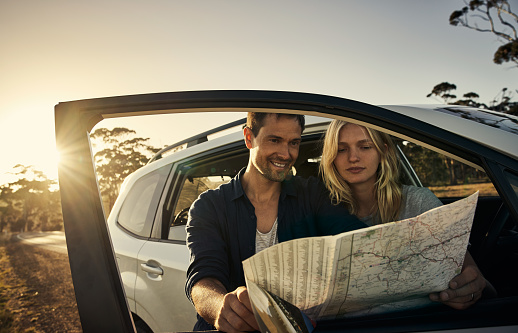 Shot of a cheerful young couple reading a map together while standing next to a open road with their car outside during the day