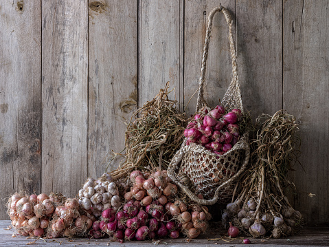 Colorful dried Thai magenta shallots hanging in a hemp string bag and other different types of shallots and garlic sitting on an old wooden table against an old wood paneled wall.