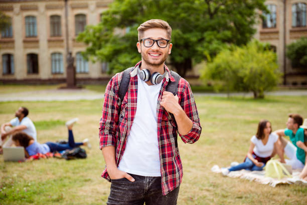 istock Young successful blond nerdy student is standing with backpack and headphones, smiles, behind are his classmates, park near campus, sunny day, carefree and enjoyable mood 936105912