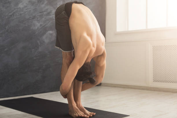Sporty man doing Standing Forward Bend posture. Guy practicing yoga, stand head to knees at gym, copy space