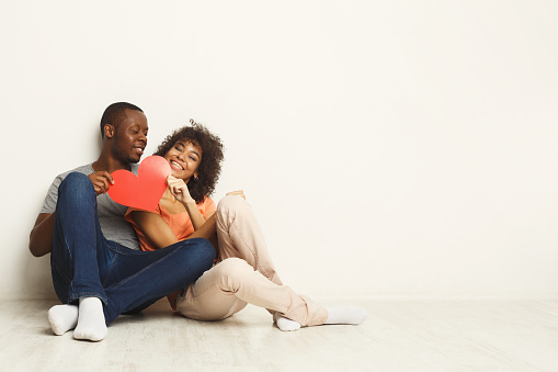 Young happy african-american couple in love holding red paper heart, sitting on floor of new flat, copy space. Family and valentine day concept, isolated