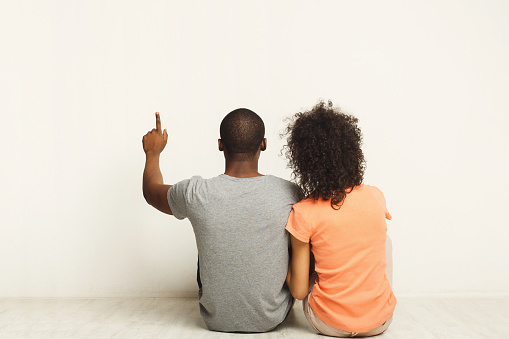 African-american family couple looking up, sitting on floor in new apartment, dreaming about future, man point upward, back view, copy space, isolated