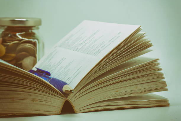 Opened book and a pen with blurred the glass jar of coin on dark background Opened book and a pen with blurred the glass jar of coin on dark background, added colour filter and classic tone effect for knowledge and education financial concept benefits of reading book stock pictures, royalty-free photos & images