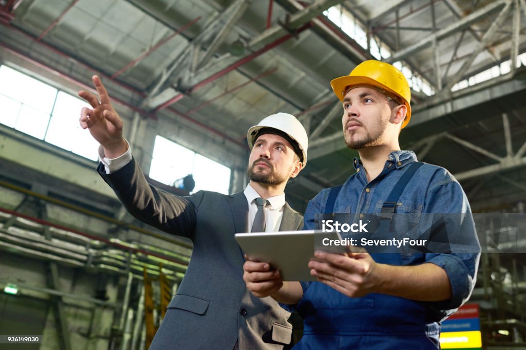 Engineers Planning at Factory Waist up portrait of handsome businessman pointing up while discussing something with factory worker holding digital tablet, copy space Manager Stock Photo