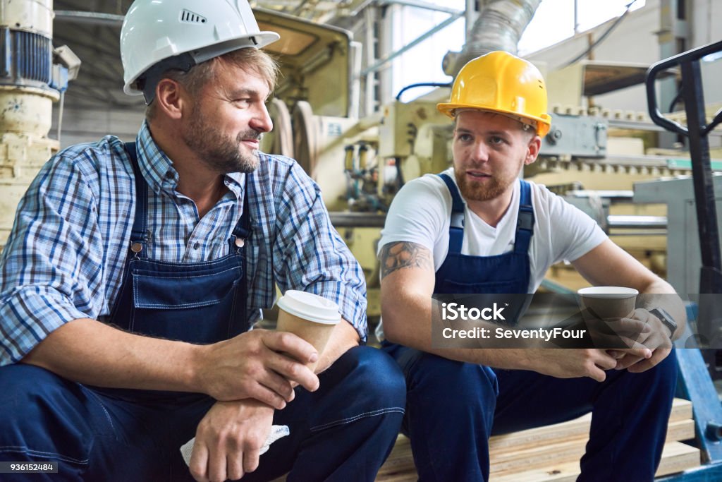 Two Workers on Coffee Break Portrait of two workers wearing hardhats taking break from work drinking coffee and resting sitting on pack of wood in workshop Construction Worker Stock Photo