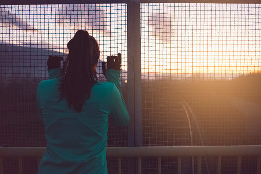 Beautiful, young female athlete standing on the bridge, looking to the highway through the fence and taking a rest after exercising.