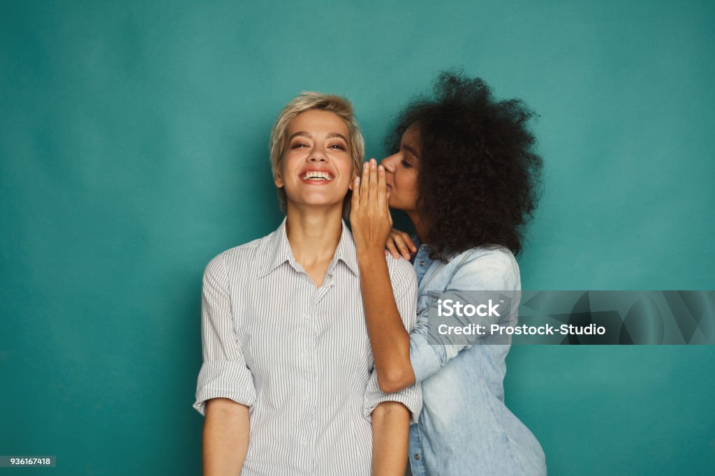 Young woman telling her friend some secrets Young woman telling her girlfriend some secret. Two women gossiping. Excited emotional girl whispering to her friend ear, turquoise studio background Whispering Stock Photo