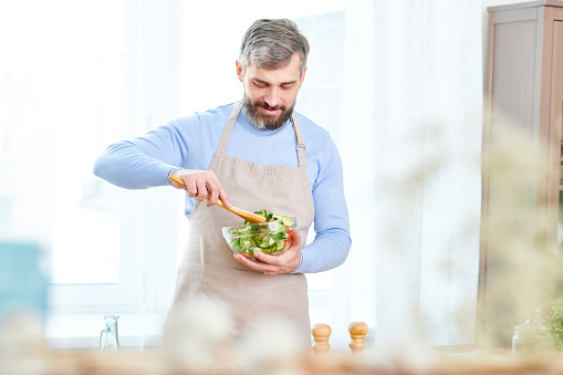Portrait of handsome bearded man mixing salad in glass bowl while cooking at home and smiling happily, copy space
