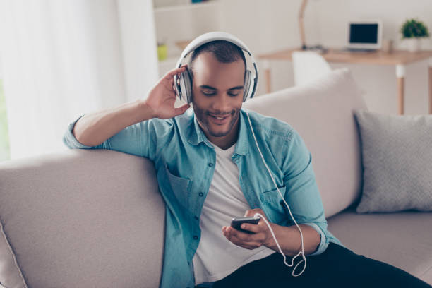 close up portrait of serene young guy enjoying listening to nice song at home, sitting on sofa, on his smart phone, wearing casual jeans outfit, smiling - young adult technology beautiful singing imagens e fotografias de stock