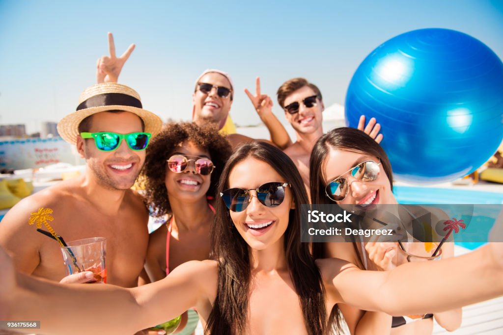 Selfie tempo! Seis amigos alegres no sol protegendo eyewear, tampões, com bebidas e desgaste da nadada diferente na moda estão posando para uma foto de selfie, está tomando aquela moça morena, irradiando sorrisos, sol - Foto de stock de Amizade royalty-free