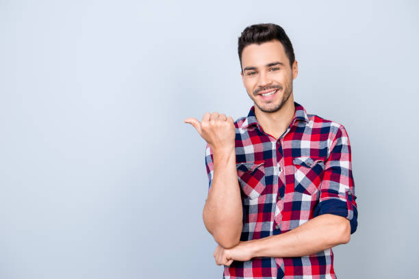istock Look here! Young cheerful man with beaming smile is pointing at te copy space with his thumb, wearing casual checkered shirt, standing on light background isolated 936176256