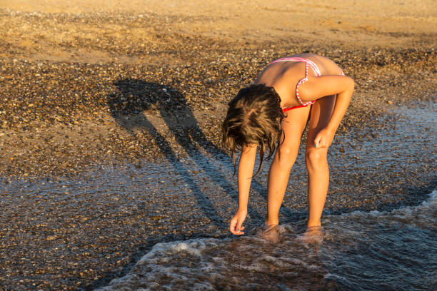 Crouched little girl picking mollusk shells on a beach in summer in Sicily, Italy