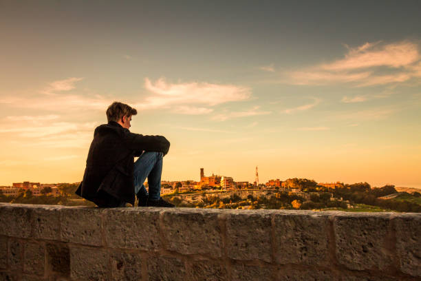 istock Lonely young man sitting on wall at sunset looking at view 936189704
