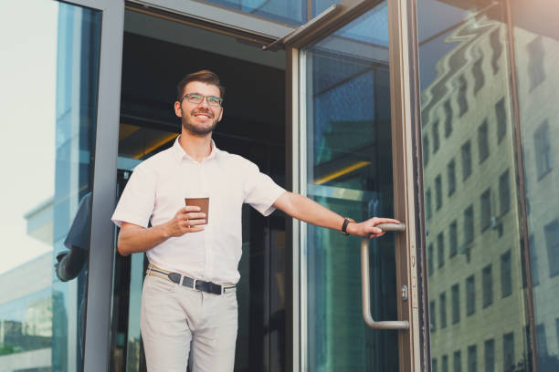 Happy smiling businessman with coffee leaving the building Confident happy young man opening the door and drinking coffee while walking out from the modern office building, copy space. Business, people and success concept office leave stock pictures, royalty-free photos & images