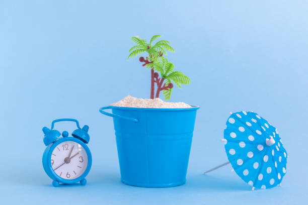 istock Alarm clock, paper cocktail parasol, can with sand and plastic palm tree isolated on blue background. Summertime vacation minimalistic still life concept. 936211808