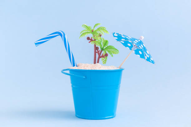 istock Drinking straw, paper cocktail umbrella, can with sand and plastic palm tree isolated on blue background. Summertime vacation minimalistic still life concept. 936212908
