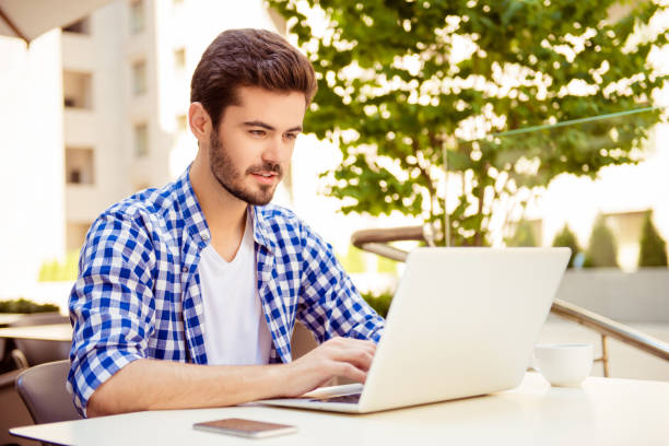 Stylish freelancer is working on his computer, sitting outdoors on a summer day, with cup of coffee, nice city view and green tree behind him