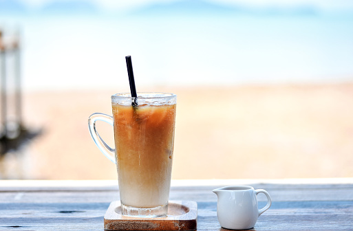 Iced cold coffee glass and sweet syrup jug at the beach bar with blurred seascape in the background.