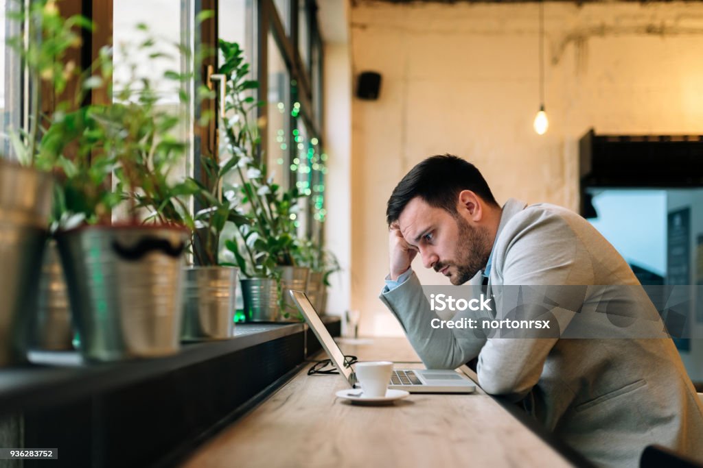 Hombre preocupado mirando la pantalla del ordenador portátil mientras está sentado en el café moderno. - Foto de stock de Confusión libre de derechos