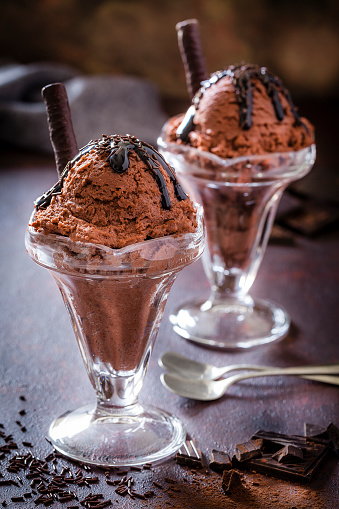 Three chocolate ice cream sundae cups shot on dark abstract table. The ice cream is covered with chocolate fudge and chocolate sprinkles. Chocolate pieces and chocolate sprinkles are scattered at the base of the cup as well as  \ntwo dessert spoons. Predominant color is brown. DSRL studio photo taken with Canon EOS 5D Mk II and Canon EF 100mm f/2.8L Macro IS USM