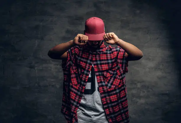 Portrait of Black bearded male dressed in a red fleece shirt and a cap over grey background.