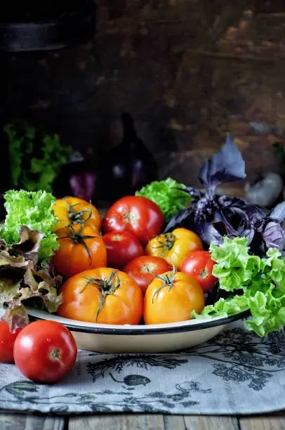 A plate of fresh vegetables and greens, on the table are tomatoes, the background is dark