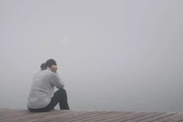 Young woman sits lonely at the edge of a wooden path of a bridge bent and sadly lost in thoughts in the fog