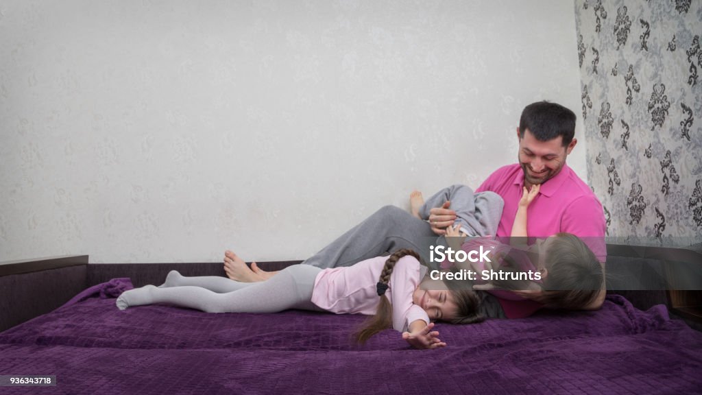 Father is tickling his two kids on bed Family is having fun. Girl is lying down and smiling. Her eyes are closed. Father holds his son on hands. Boy pulls his hands to father's face. Adult Stock Photo