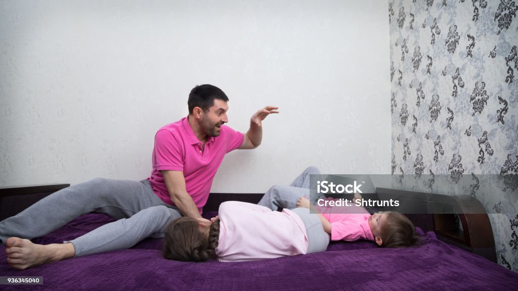 Father and his two kids are playing on bed Family is having fun. Girl is lying on one side. Father is sitting and scaring his son. Boy is lying on the back and laughing. Main colors are white, pink, purple, gray and black. Black Color Stock Photo