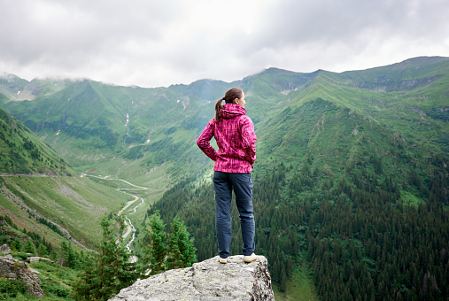 Young female tourist standing on rock edge among magnificent green mountains with grassy slopes and winding roads near Transfagarashan road in Romania. Woman climber trees beautiful view scenery