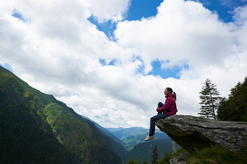Profile of the girl sitting on the rock, dangling her legs in the abyss against the backdrop of green mountains with forests and clouds above them through which the blue sky is visible.