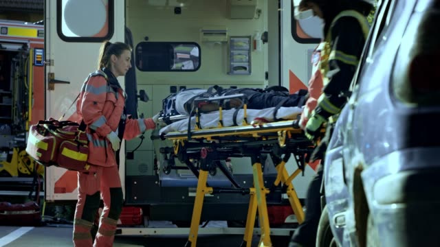Two male paramedics loading the injured person into the ambulance and the female doctor accompanies the patient