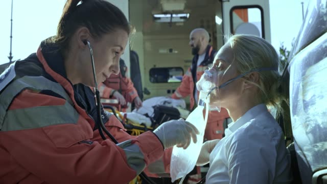 Female paramedic talking to the injured woman sitting on the ground at the scene of a car accident and breathing with the oxygen mask