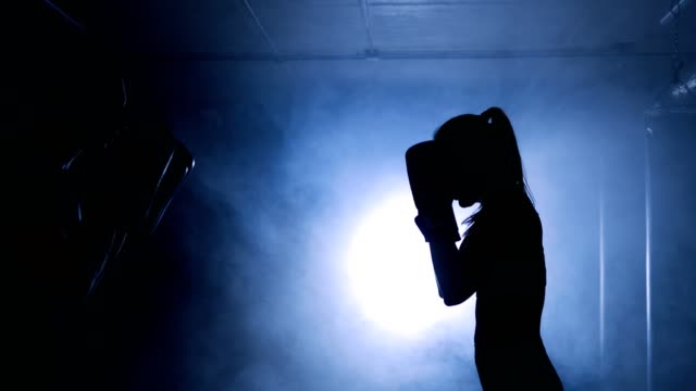 Young woman training with a coach in a boxing club in a smoky gym. Silhouette