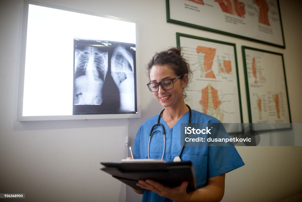 Portrait of charming beautiful middle-aged professional female doctor writing on clipboard in front of x-ray panel in a hospital office. X-ray Image Stock Photo