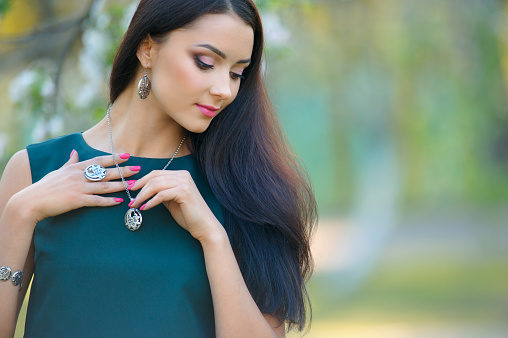 Beautiful young woman in red sleeveless top wearing ruby jewelry, standing in public park and smiling looking away, beauty and fashion industry