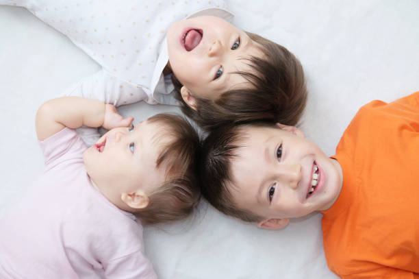 crianças felizes, três crianças rindo diferentes idades mentindo, retrato de menino, menina e bebê menina, felicidade na infância dos irmãos, vivendo em família grande - sibling baby three people baby girls - fotografias e filmes do acervo