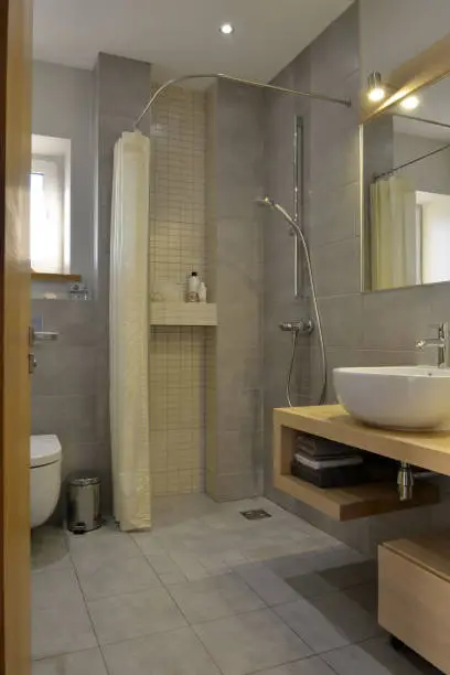 Grey stone pattern tiled contemporary bathroom interior design with minimal natural wood furniture details. Shower, washbasin, mirror, toilet