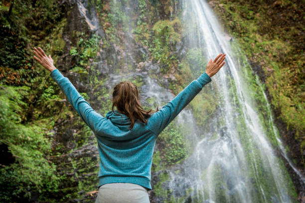 istock Young woman with arms wide open standing near waterfall 936407844