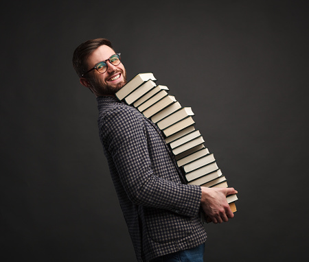 Side view of content male student carrying huge stack of library books smiling at camera.