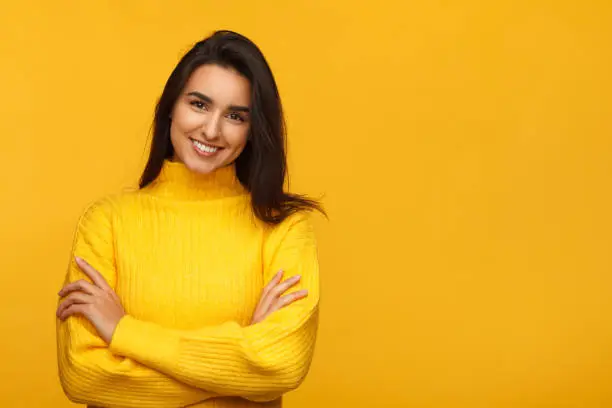 Young beautiful brunette in yellow sweater standing with hands crossed on yellow.